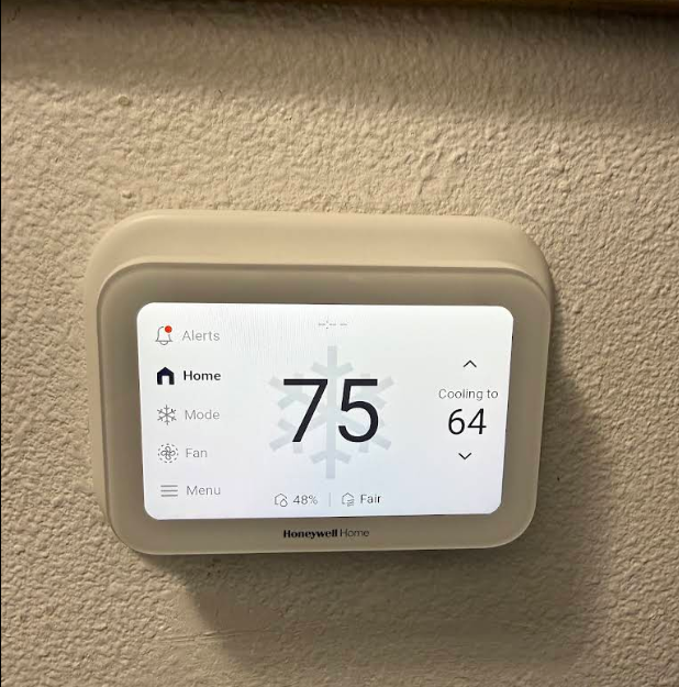 An image showing a white Honeywell Home smart thermostat mounted on a textured, beige wall. The thermostat's screen is lit, displaying the current indoor temperature as 75 degrees in large black numbers with a faint snowflake icon behind them. On the right, it shows "Cooling to 64" with up and down arrows. The left side of the screen features a menu with options for Alerts, Home, Mode, Fan, and Menu. At the bottom, it indicates 48% humidity and "Fair" air quality.