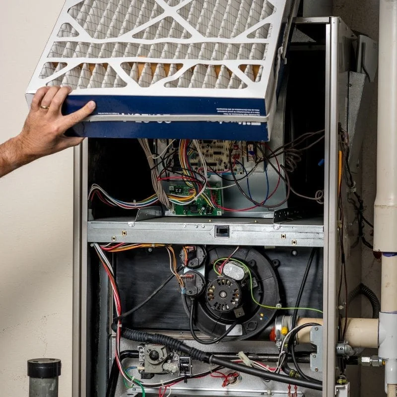 An eye-level shot captures the internal components of a silver-colored furnace against a white wall. At the top, a hand with a silver ring on the ring finger lifts a thick, white, pleated air filter. Below the filter, various electrical components, including green and tan circuit boards and a multitude of colorful wires (white, red, yellow, and blue), are visible. In the bottom half of the furnace, a black circular fan motor is mounted, surrounded by more wires and a black, corrugated hose. To the right of the furnace, a white PVC pipe runs vertically.