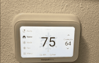 An image showing a white Honeywell Home smart thermostat mounted on a textured, beige wall. The thermostat's screen is lit, displaying the current indoor temperature as 75 degrees in large black numbers with a faint snowflake icon behind them. On the right, it shows "Cooling to 64" with up and down arrows. The left side of the screen features a menu with options for Alerts, Home, Mode, Fan, and Menu. At the bottom, it indicates 48% humidity and "Fair" air quality.