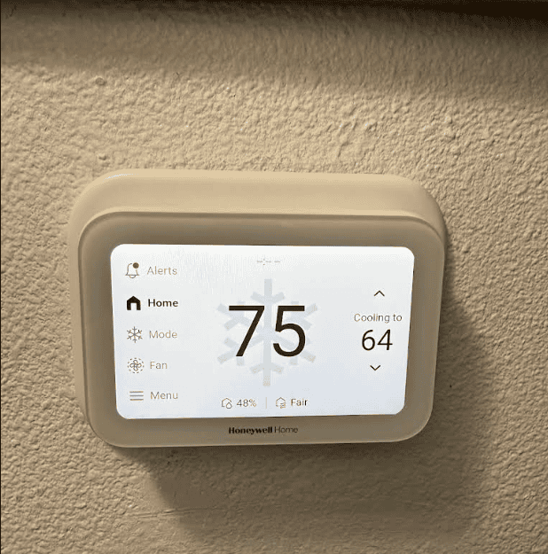 How Do I Calculate the Cooling Capacity Needed for My Home? An image showing a white Honeywell Home smart thermostat mounted on a textured, beige wall. The thermostat's screen is lit, displaying the current indoor temperature as 75 degrees in large black numbers with a faint snowflake icon behind them. On the right, it shows "Cooling to 64" with up and down arrows. The left side of the screen features a menu with options for Alerts, Home, Mode, Fan, and Menu. At the bottom, it indicates 48% humidity and "Fair" air quality.