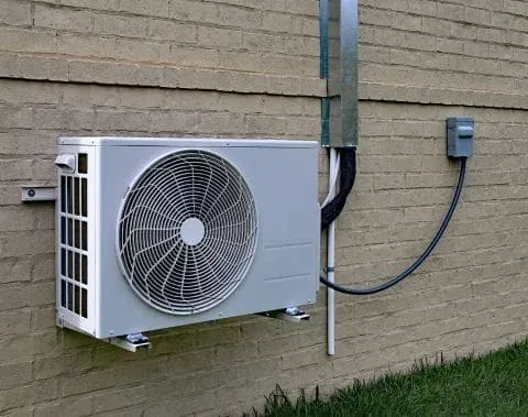 A close-up shot of a gray, rectangular air conditioning unit, specifically a ductless mini-split system, mounted on a tan brick wall. The unit has a large circular fan grille on the front and is connected to the building by wiring and pipes.