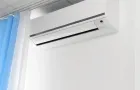 The image features a sleek, white air conditioning unit installed high on a white wall, near a suspended ceiling and blue and white curtains.