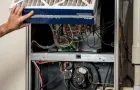 An eye-level shot captures the internal components of a silver-colored furnace against a white wall. At the top, a hand with a silver ring on the ring finger lifts a thick, white, pleated air filter. Below the filter, various electrical components, including green and tan circuit boards and a multitude of colorful wires (white, red, yellow, and blue), are visible. In the bottom half of the furnace, a black circular fan motor is mounted, surrounded by more wires and a black, corrugated hose. To the right of the furnace, a white PVC pipe runs vertically.